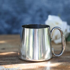 May include: A polished silver-toned metal mug with a smooth, slightly tapered cylindrical body and a curved handle attached to the side. The surface of the mug is reflective, showing subtle signs of wear and small dark spots across the exterior.