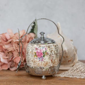 May include: A vintage ceramic sugar bowl with a floral design and a silver lid and handle. The bowl is on a silver stand with three feet.