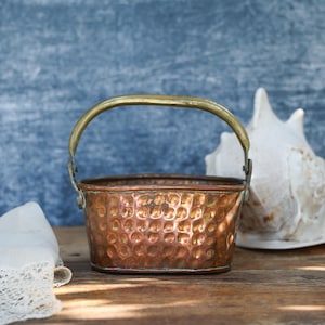 May include: A small, copper-colored bucket with a brass handle. The bucket has a textured, dimpled surface. A white seashell and a piece of lace are in the background. The bucket sits on a wooden surface.