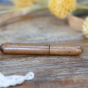 May include: A small, brown, cylindrical object with a split in the middle, possibly a pencil holder or similar item. The object is made of a light brown material with darker brown speckles. The background is a wooden surface with dried yellow flowers.