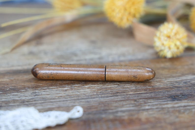 May include: A small, brown, cylindrical object with a split in the middle, possibly a pencil holder or similar item. The object is made of a light brown material with darker brown speckles. The background is a wooden surface with dried yellow flowers.