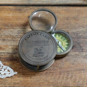 May include: A round brass map reader compass with a magnifying glass lid. The lid features the text CAPTAIN CABIN, MAP READER COMPASS WITH MAGNIFYING GLASS, It is very useful in navigation, Science, and Please lock the compass after use, with a ship icon.