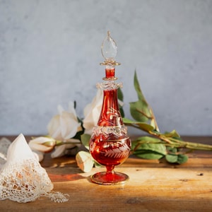 Könnte beinhalten: Rote Glasparfümflasche mit goldfarbenem Verschluss und Akzenten. Die Flasche hat ein geätztes Design und ist mit roter Flüssigkeit gefüllt. Sie steht auf einer Holzoberfläche mit weißen Blumen und Grün.