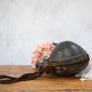 May include: A dark gray ceramic water bottle with a rope handle. The bottle is sitting on a wooden surface with a pink flower in the background.
