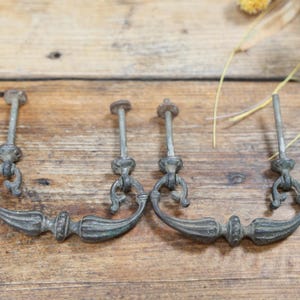 May include: Two matching metal drawer pulls with a weathered, dark grey finish. Each pull features a curved, ornate handle attached to two vertical mounting posts with circular bases. The design includes intricate, symmetrical detailing.