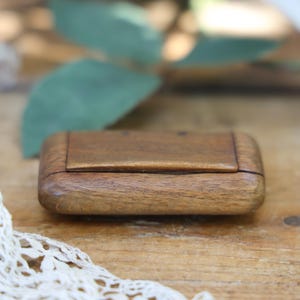 May include: A small, rectangular wooden box with a smooth, polished finish and rounded edges. The box features a hinged lid that sits flush with the main body, showcasing a natural brown wood grain pattern throughout the entire exterior surface.
