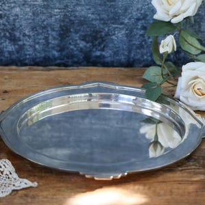 May include: A polished silver-toned metal serving tray with a smooth, reflective circular centre and a wide, flat rim featuring subtle geometric notched edges. The tray has a metallic finish that reflects the surrounding light and nearby objects.