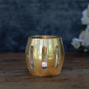 May include: A polished gold-toned metal candle holder with a barrel-like shape and vertical cutouts. The cylindrical vessel features a smooth, reflective surface and contains a small, lit white tea light candle visible through the openings.