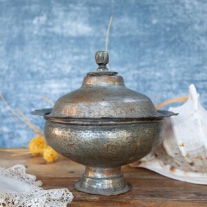 May include: An antique, lidded metal bowl with a hammered texture, set on a pedestal base. The bowl has a tarnished, aged appearance. A large seashell and lace are in the background, on a wooden surface, against a blue backdrop.