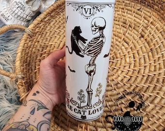 May include: White ceramic tumbler with a black and white illustration of a skeleton holding a black cat. The text "VI The Cat Lover" is printed on the tumbler.