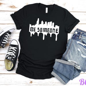 May include: Black t-shirt with the white text "BE SOMEONE" in a cityscape design with dripping paint effect. The shirt is paired with black Converse sneakers and blue denim jeans.