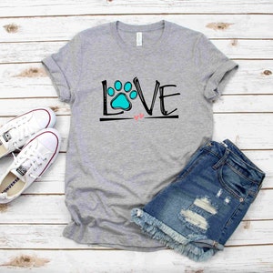 May include: A gray t-shirt with the word "LOVE" written in black with a blue paw print in the "O". Two small pink hearts are below the word. The t-shirt is on a white wooden background with a pair of white sneakers and blue denim shorts.