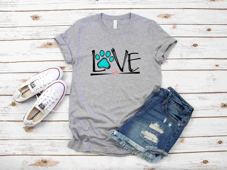 May include: A gray t-shirt with the word "LOVE" written in black with a blue paw print in the "O". Two small pink hearts are below the word. The t-shirt is on a white wooden background with a pair of white sneakers and blue denim shorts.