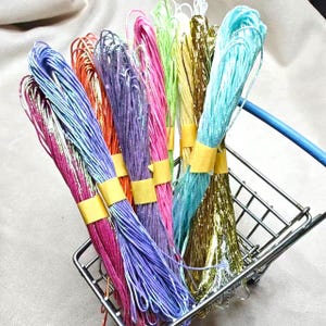 Puede incluir: Una variedad de coloridos paquetes de hilo artesanal, que incluyen tonos de rosa, morado, naranja, azul y dorado. Cada paquete está asegurado con cinta adhesiva amarilla y se presenta en un pequeño carrito de compras de metal. Los hilos parecen estar hechos de un material brillante y metálico.