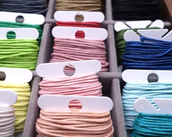 May include: A close-up of a craft organizer filled with spools of colorful thread. The threads are in various shades, including pink, green, blue, and yellow. Each spool is neatly wound and held in place with a white plastic holder. The organizer is made of a gray material.