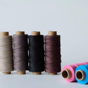 May include: Six cylindrical spools of thread arranged in a row. Four spools stand vertically in beige, grey, black, and dark brown, while two spools lie horizontally in bright pink and vibrant blue. Each spool features a cardboard core center.