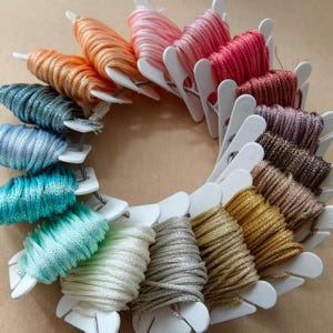 May include: A circular arrangement of embroidery floss bobbins featuring a spectrum of metallic and solid colors including orange, pink, red, teal, blue, gold, silver, and brown. Each white plastic bobbin holds a neatly wound length of thread.