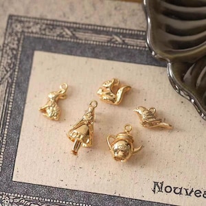 May include: Five gold-coloured charms in various shapes: a rabbit, a girl, a squirrel, a bird, and a teapot. The charms are small and detailed, with a loop at the top for attaching to jewellery or other crafts. The background is a light beige colour.
