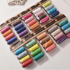 Könnte beinhalten: Mehrere Packungen Hanf-Garn in verschiedenen Farben, darunter Rosa, Blau, Grün und Braun. Jede Packung enthält mehrere Garnrollen, mit dem Aufdruck "HEMP THREAD" auf der Verpackung.