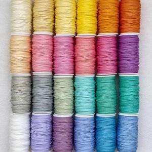 Può includere: Una collezione di rocchetti di filo colorati, disposti in file ordinate. I colori del filo includono sfumature di giallo, rosa, verde, blu e arancione. Le bobine sono su bobine di plastica bianca e il filo sembra essere ben avvolto.