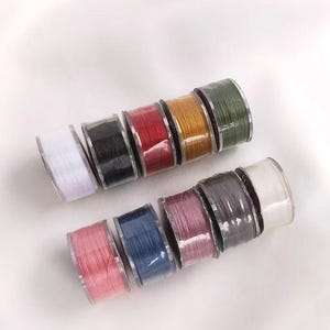 May include: A collection of spools of thread in various colors, including white, black, red, green, blue, and pink. The spools are arranged in a row, with the white spool at the end of the row.