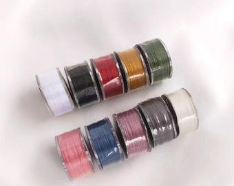 Könnte beinhalten: Eine Sammlung von Garnrollen in verschiedenen Farben, darunter Weiß, Schwarz, Rot, Grün, Blau und Pink. Die Rollen sind in einer Reihe angeordnet, wobei die weiße Rolle am Ende der Reihe liegt.