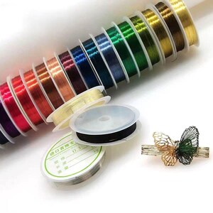 Peut inclure: Plusieurs bobines de fil de perlage coloré, notamment rose, rouge, orange et vert. Plusieurs bobines de fil noir et argent sont également visibles, ainsi qu'une barrette à cheveux papillon dorée et verte.