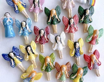 May include: A collection of 18 small ceramic fairy figurines with painted wings and dresses in various colors, including blue, green, red, pink, yellow, and white. The fairies are arranged on a white surface.