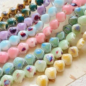 May include: Close-up of strands of colorful beads in various shapes and colors. The beads include round, shell, and square shapes in shades of pink, blue, green, yellow, and brown. Some beads have a marbled or speckled appearance.
