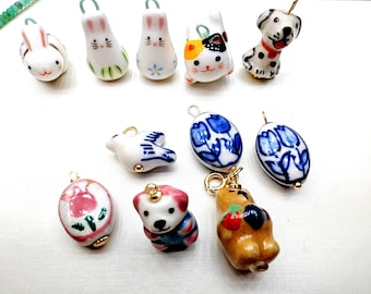 May include: A collection of ceramic charms featuring various animal and floral designs. The charms are white with colorful details, including a blue bird, a pink flower, a brown bear, a white rabbit, a white cat, a white dog, and a blue tulip.