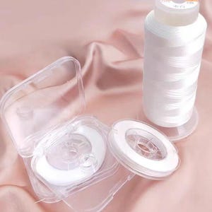 Op de afbeelding: Een close-up van witte naaigaren spoelen en doorzichtige plastic doosjes. Eén grote spoel staat rechtop, terwijl kleinere spoelen in open doosjes zitten. De achtergrond is een zachte, roze stof.
