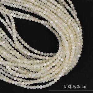 Puede incluir: Una hebra de cuentas de concha iridiscente blanca, de aproximadamente 3 mm de diámetro. Las cuentas están enhebradas en un hilo transparente y están dispuestas en un patrón circular.