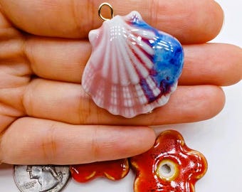 May include: A collection of ceramic pendants and charms. The main pendant is a seashell shape with pink, white, and blue glaze. Additional charms include a red flower and a red bow. A gold-colored loop is attached to the seashell pendant.