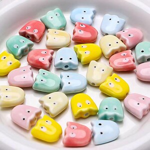Op de afbeelding: Een verzameling keramische kralen in de vorm van een spook in verschillende pastelkleuren, waaronder roze, blauw, geel en groen. Elke kraal heeft een klein gaatje om te rijgen.
