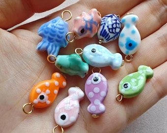 May include: A collection of colorful ceramic fish charms in various shapes and sizes. The charms are in shades of blue, pink, orange, green, and white, with gold-colored loops for attaching to jewelry or other crafts. Some have polka dots.