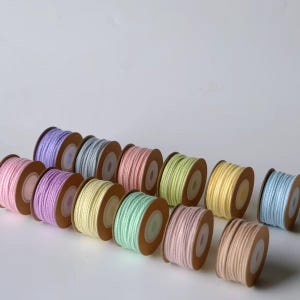 May include: A collection of twelve small spools of cord in various pastel colours, including shades of pink, purple, blue, yellow, and green. Each spool features a brown cardboard core and is neatly wound with smooth, thin, solid-coloured cord.