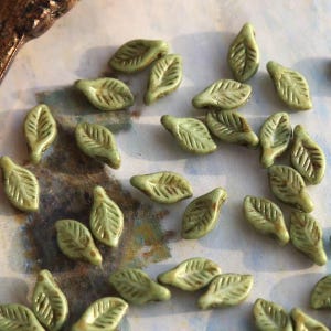 Può includere: Una collezione di piccole perline a forma di foglia verde oliva. Ogni perlina presenta venature dettagliate e una finitura leggermente lucida. Le perline sono sparse su una superficie, suggerendo il loro utilizzo in progetti di gioielleria o artigianato.