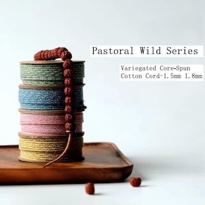 Puede incluir: Una pila de cuatro carretes de cordón de algodón hilado con núcleo jaspeado en verde, azul, rosa y amarillo. Un hilo de cuentas marrones texturizadas cuelga por el lado. El texto dice: Pastoral Wild Series, Variegated Core-Spun Cotton Cord-1.5mm 1.8mm.