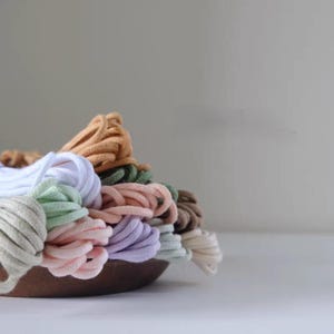 Puede incluir: Una colección de cordones de tela enrollados en varios colores pastel y neutros, incluyendo blanco, rosa claro, lavanda, verde salvia, beige y marrón, dispuestos en un cuenco de madera redondeado. Los cordones tienen una textura lisa y redondeada.