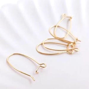 Puede incluir: Ganchos de pendientes dorados de varias formas y tamaños sobre un fondo blanco. Los ganchos están hechos de metal fino y curvado, con un bucle en el extremo para sujetar los pendientes. El diseño es simple y elegante, adecuado para la fabricación de joyas.