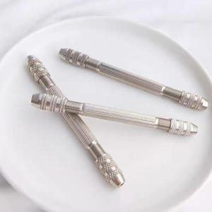 Op de afbeelding: Drie zilveren handboormachines van metaal met een getextureerde grip en een puntige punt. De boormachines liggen op een wit bord.