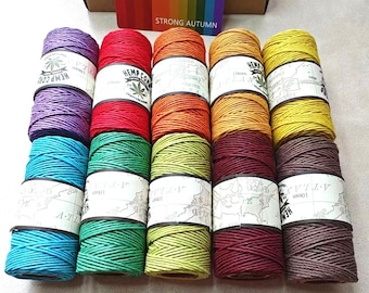 Peut inclure: Plusieurs bobines de fil de chanvre colorées, dans des tons de violet, rouge, orange, jaune, turquoise, vert et marron. Chaque bobine est étiquetée avec du texte et un logo de feuille de cannabis. La boîte en arrière-plan présente un dégradé arc-en-ciel et le texte "STRONG AUTUMN".