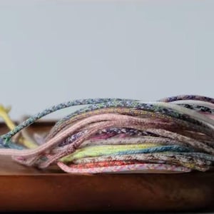 May include: A collection of thin, braided cord bracelets in a variety of pastel and vibrant colours including pink, yellow, blue, and green. The cords are woven with a textured pattern and are layered together in a small, rounded pile.