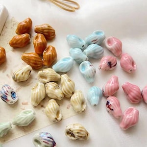 Può includere: Una collezione di perline in ceramica in vari colori, tra cui arancione, azzurro, rosa e crema. Le perline hanno una forma a conchiglia con superfici strutturate e sono sparse su una superficie bianca. Alcune perline presentano dettagli dipinti.