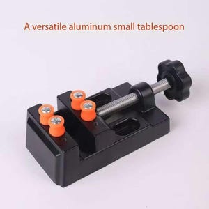 Puede incluir: Un tornillo de banco de metal negro con un tornillo plateado y una perilla negra. El tornillo de banco tiene cuatro almohadillas de goma naranja en las mordazas.