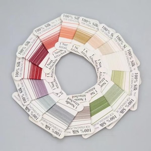 Pode incluir: Uma disposição circular de vários cartões de fio 100% seda para enfiar pérolas e contas. Cada cartão apresenta uma cor de fio diferente, incluindo vermelho, rosa, verde, cinzento e branco, com o texto 100% Silk e 1 Needle Attached.