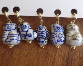 May include: Five ceramic animal earrings with gold-colored accents. The earrings feature a cat, dog, fish, owl, and another cat, all painted with blue and white designs. The earrings are hanging from a white card.
