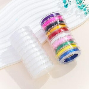 Puede incluir: Dos pilas cilíndricas de cordón fino y flexible. Una pila contiene cordón transparente e incoloro. La otra pila presenta una variedad de colores vibrantes, incluyendo cordón morado, rosa, negro, naranja, rojo, blanco, rosa claro, verde, amarillo y azul.
