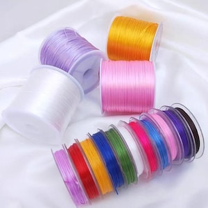 May include: Assortment of colorful elastic cords in various colors, including white, pink, yellow, purple, red, orange, blue, green, and black. The cords are wound around spools and are ready for crafting projects.