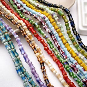 Puede incluir: Una colección de cuentas coloridas en forma de hueso en varios colores, incluyendo rojo, verde, azul, amarillo y morado. Las cuentas están ensartadas, creando un patrón vibrante y texturizado. Estas cuentas son adecuadas para la fabricación de joyas y proyectos de artesanía.